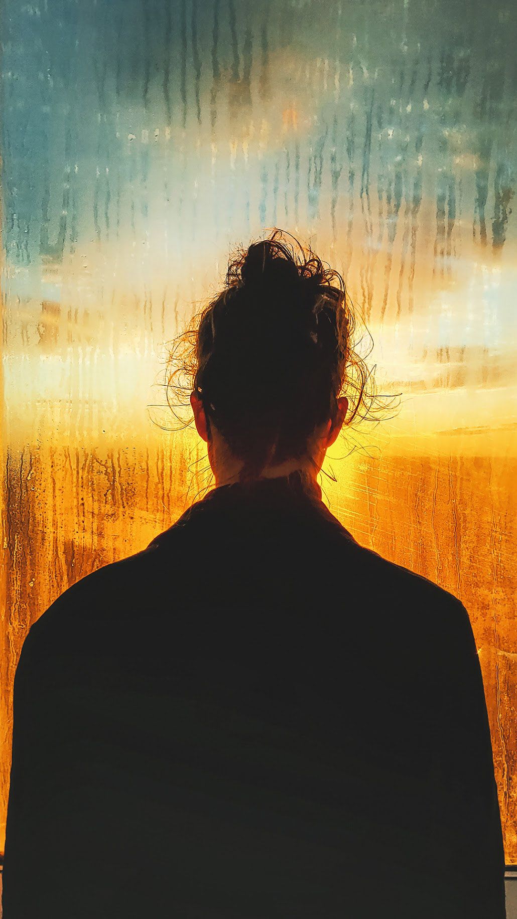 A person seen from behind, hair in a loose bun, standing in silhouette against a rain-streaked window glowing amber and teal at sunset.