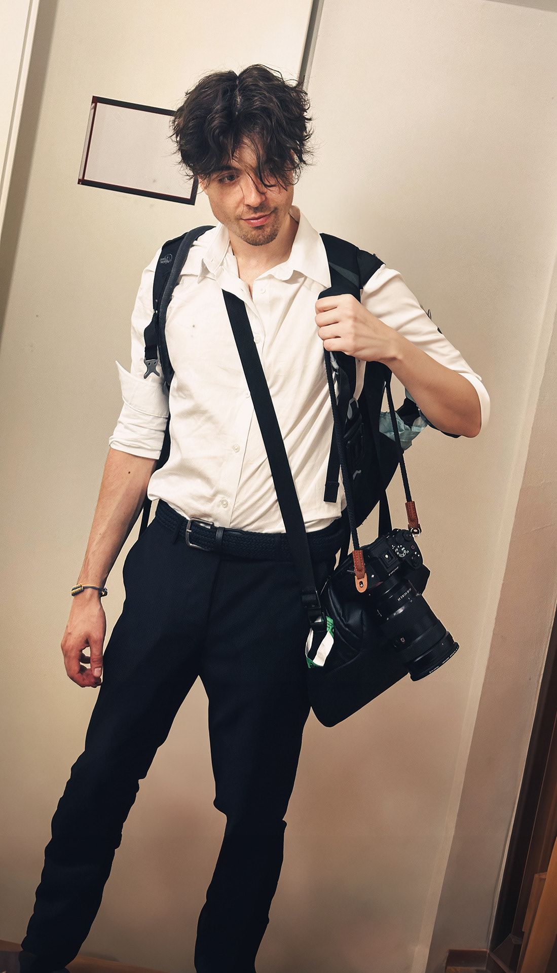 Yan Stroeb in a white shirt and black trousers, backpack on, camera with a long lens hanging at the hip, glancing down with a slight smile.
